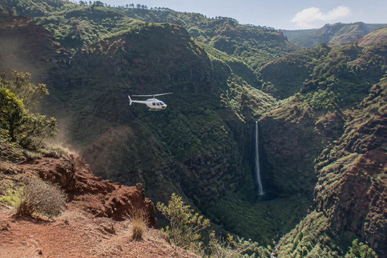 A helicopter flying over deep green canyon cliffs and a thin waterfall on Kauai