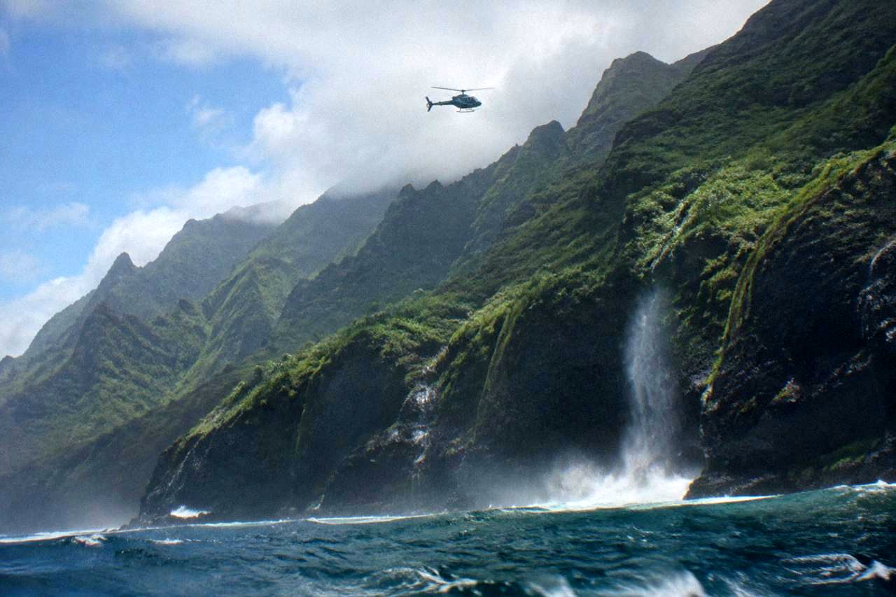 A helicopter hovering over green Kauai cliffs above the ocean