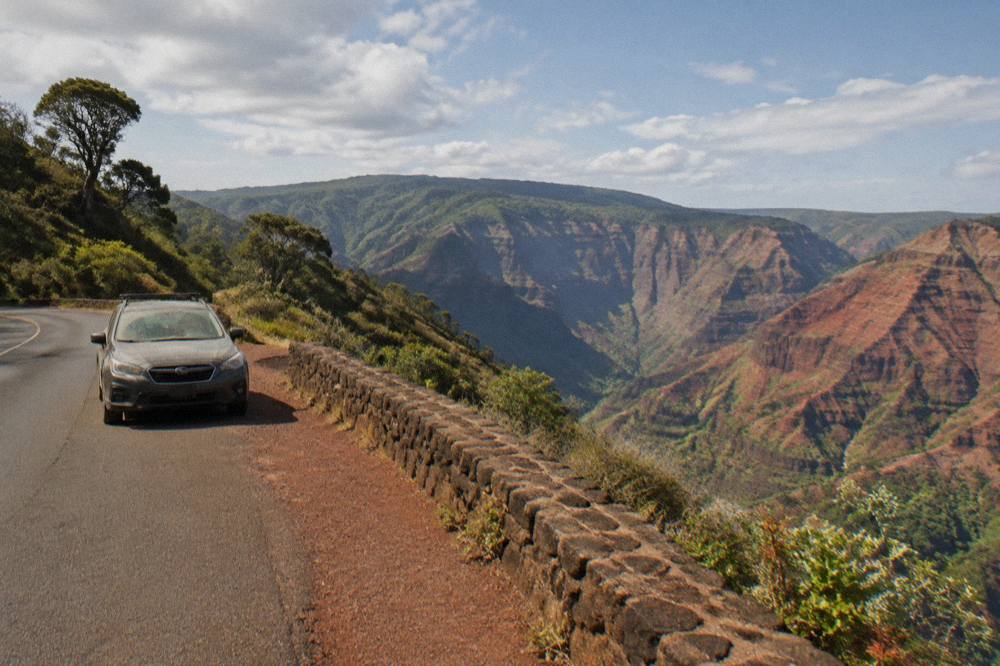 A paved mountain road curving along a red rock canyon rim with green ridges beyond