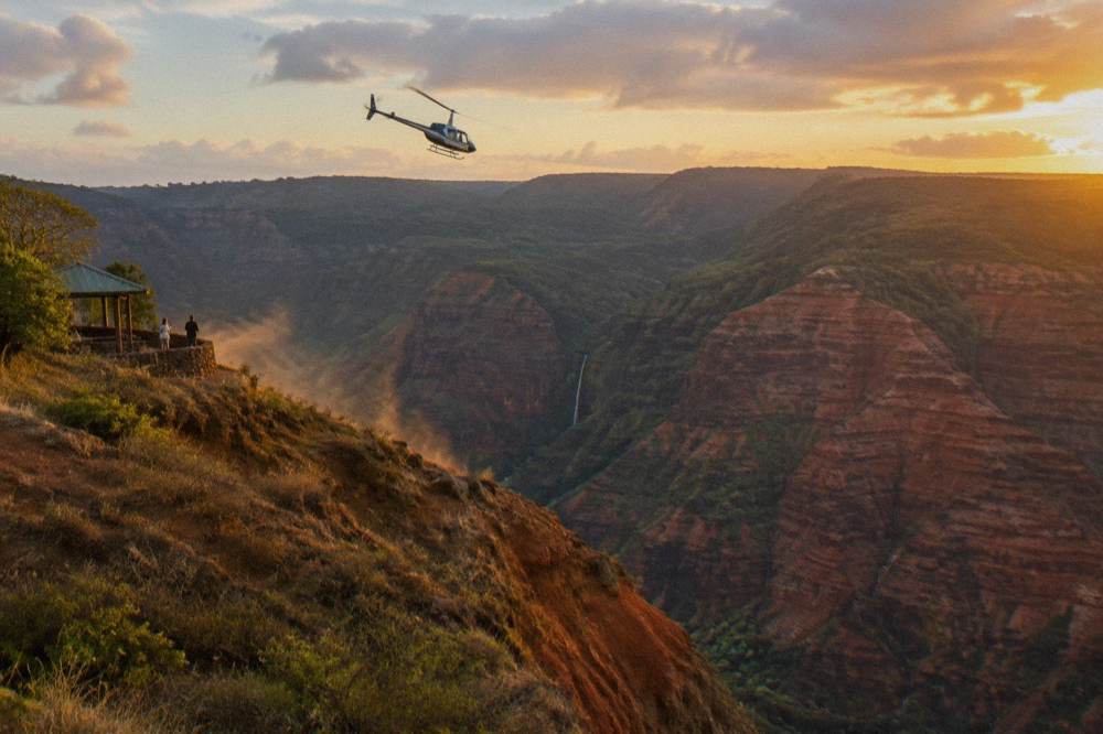 A helicopter flying above deep red canyon walls and green valley ridges
