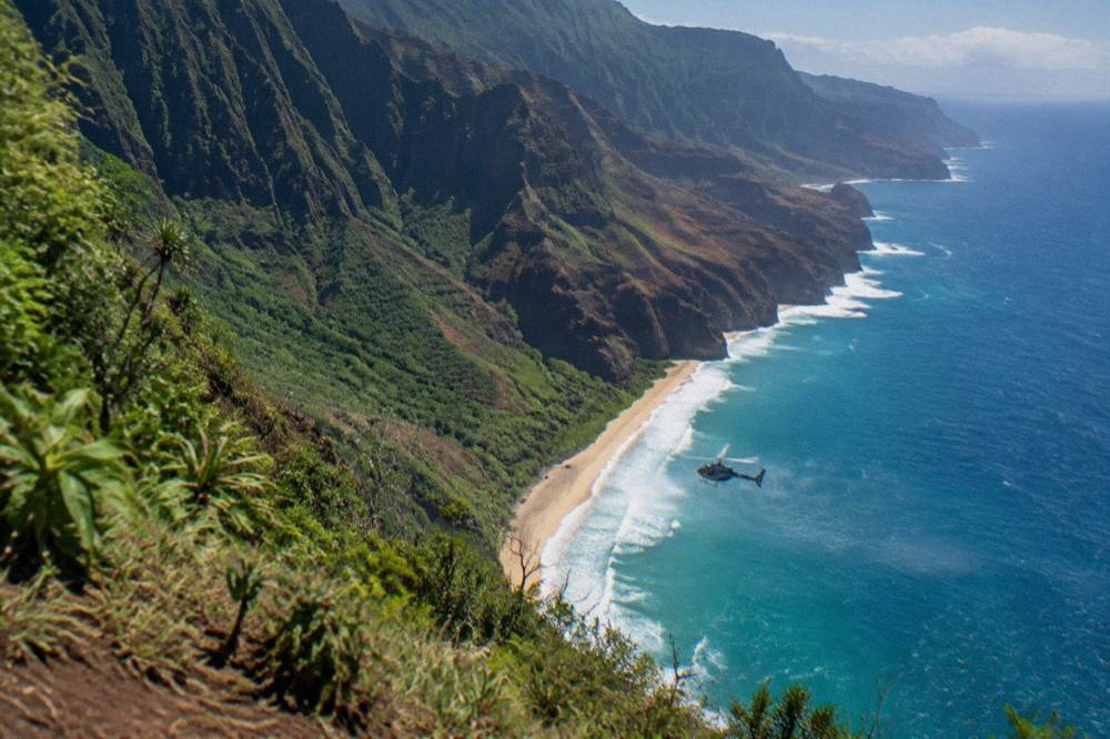 A helicopter flying above a coastline of tall green cliffs and breaking surf