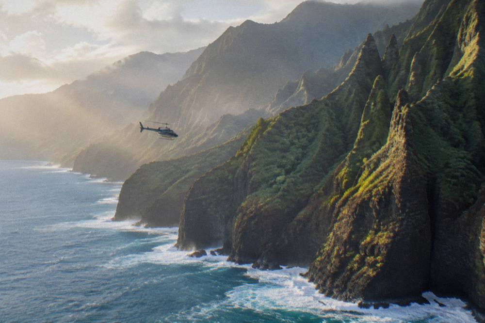 A helicopter flying low over the Na Pali Coast, with sheer green cliffs and blue ocean below