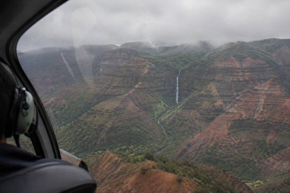 A helicopter cockpit window with a view of Waimea Canyon and a waterfall below, no people visible