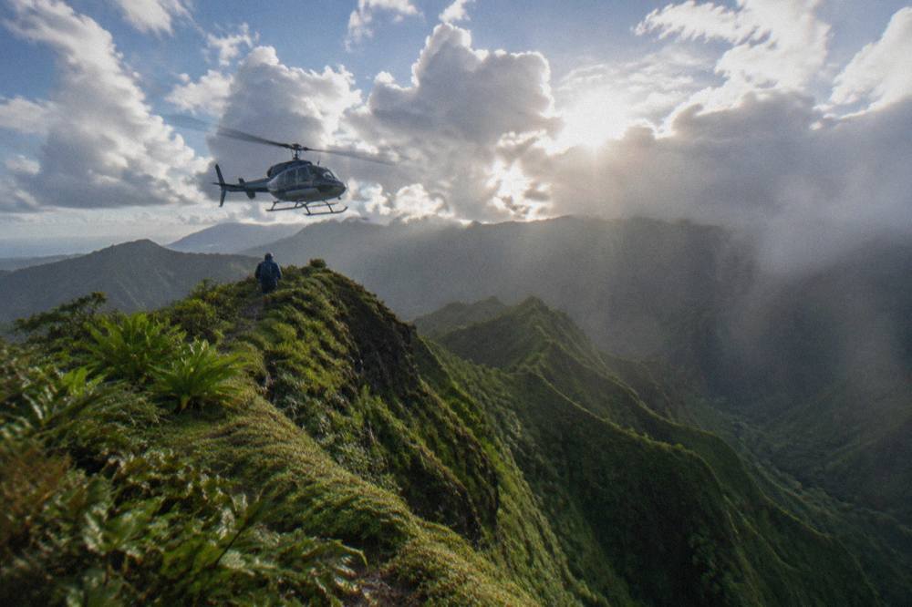 A helicopter hovering above a ridge line above a deep green canyon under scattered clouds
