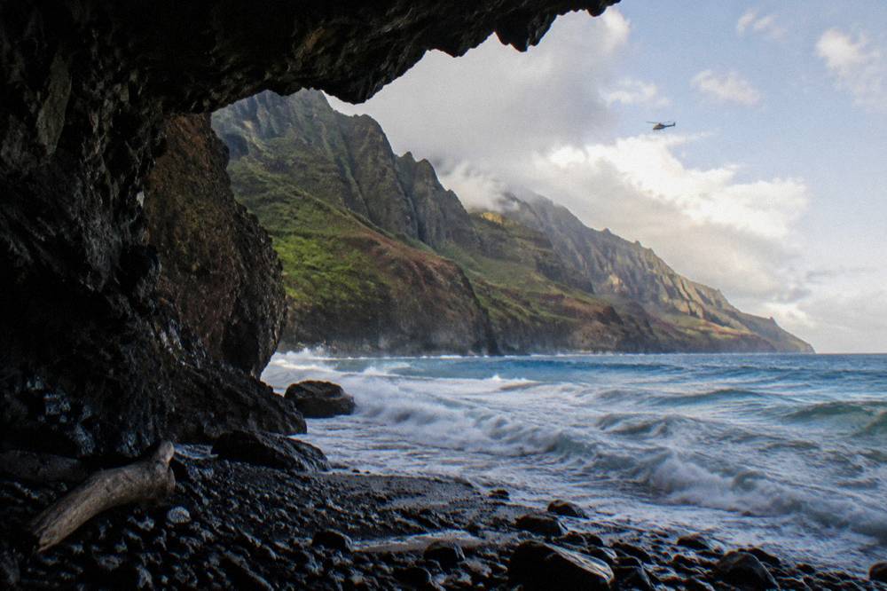 A small helicopter flying along a coastline of tall sea cliffs above breaking surf