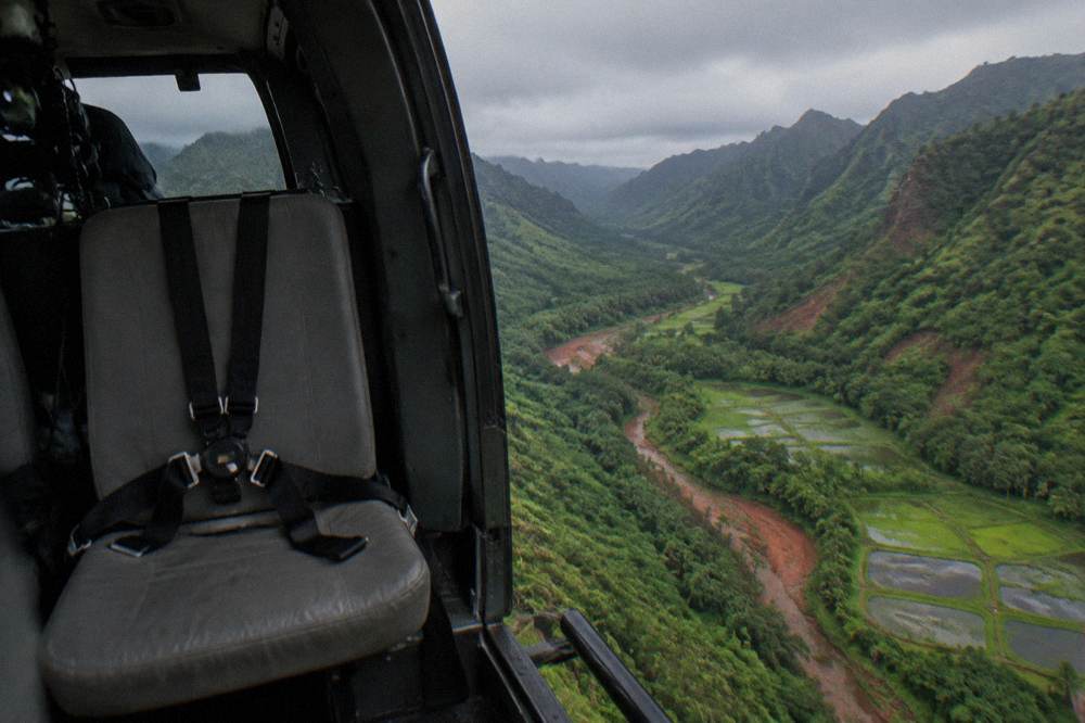 An open-door helicopter cabin showing an empty seat with a five-point harness above a forested valley