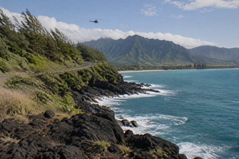 A helicopter flying above a winding coastal road along green cliffs