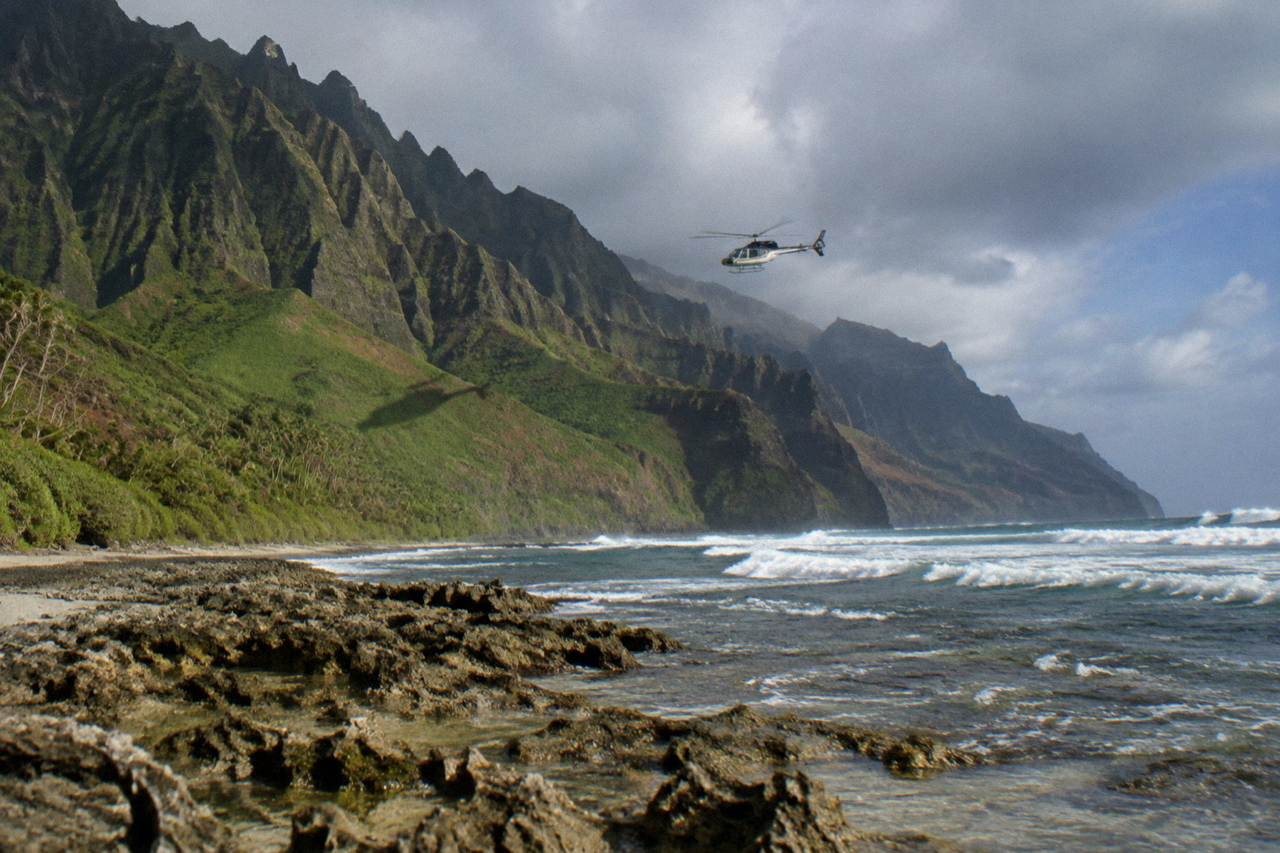 A tour helicopter in flight above green Kauai cliffs and the ocean