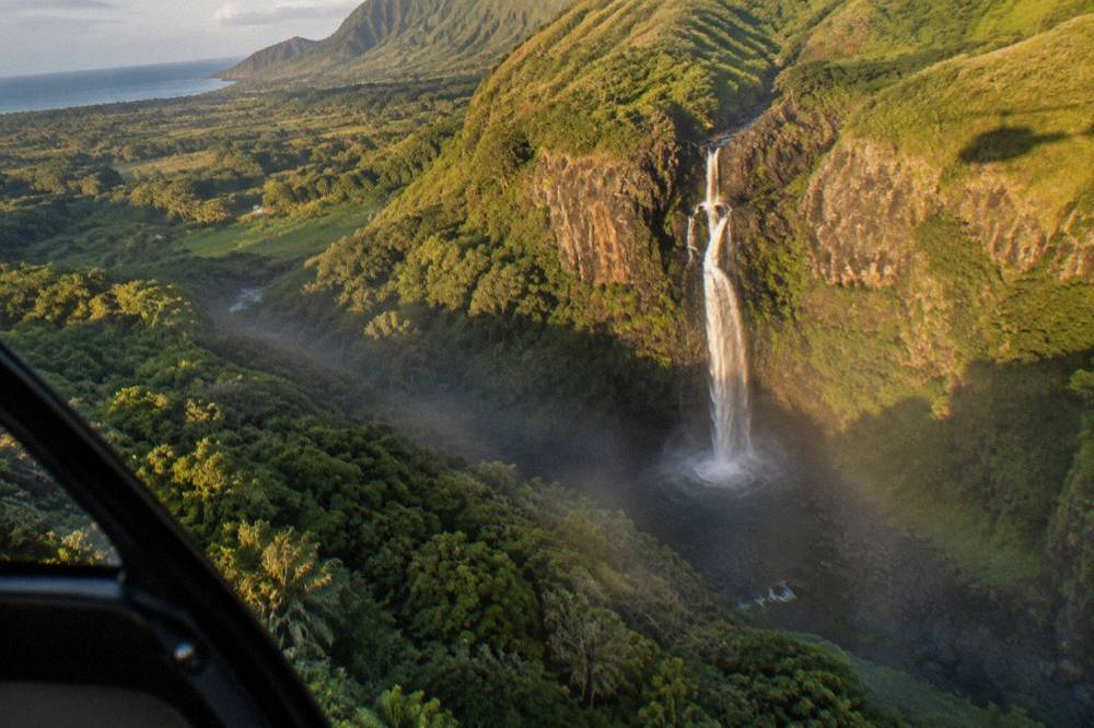 An aerial view of a tall waterfall pouring down a dark cliff into a dense green valley