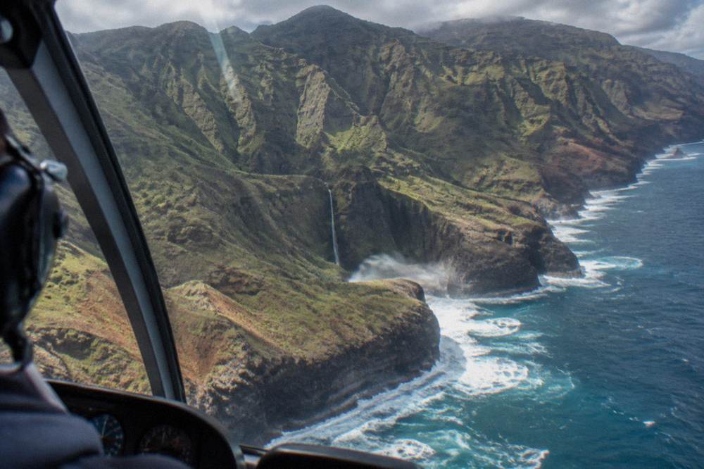 A helicopter flying along a rugged coastline with tall sea cliffs and breaking surf below