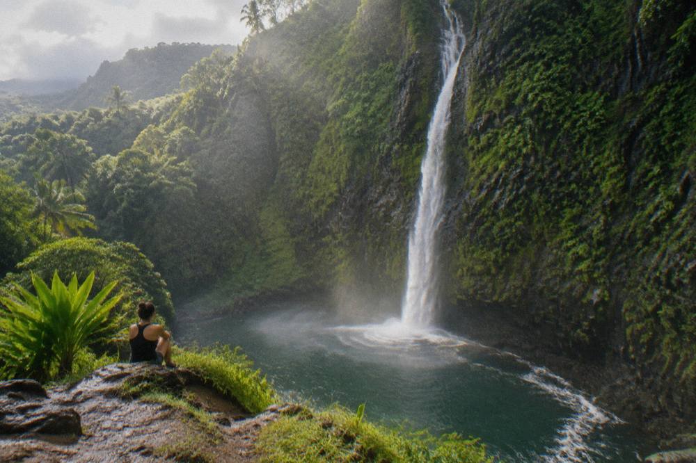A tall thin waterfall pouring down a fluted green cliff into a jungle valley