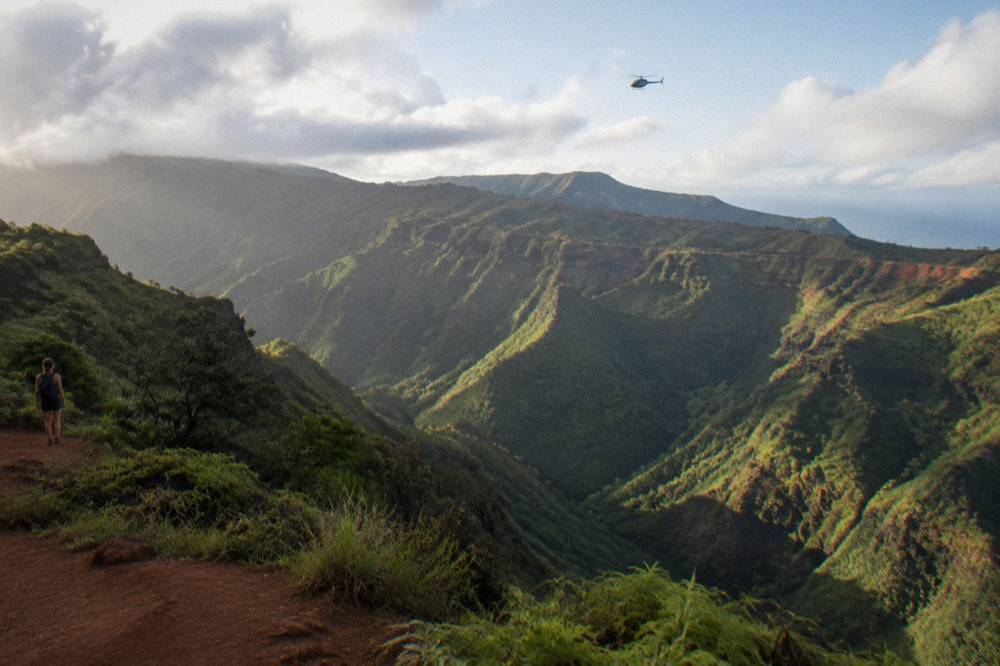 A helicopter flying above a deep green canyon with layered ridges under scattered clouds