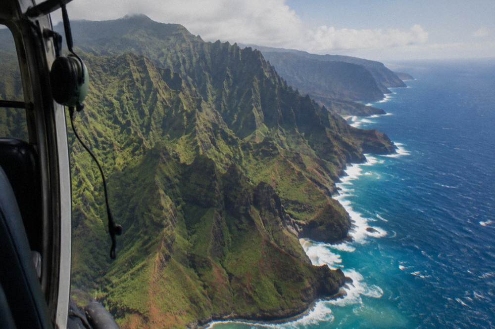 An open helicopter door view of a jagged coastline with green cliffs meeting the ocean