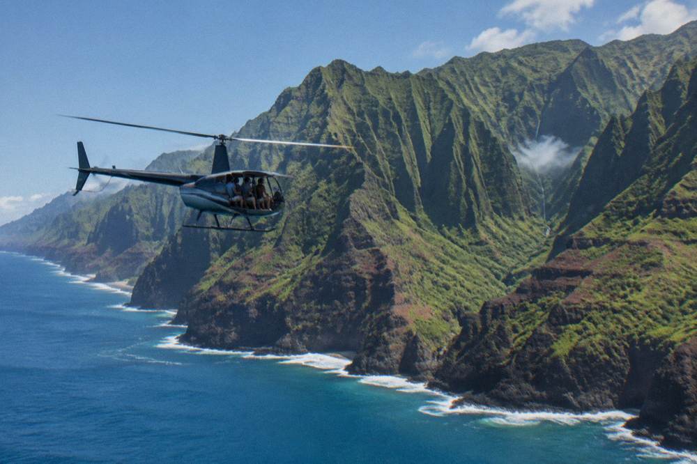 A coastline of tall sea cliffs with a helicopter flying parallel to the ridges