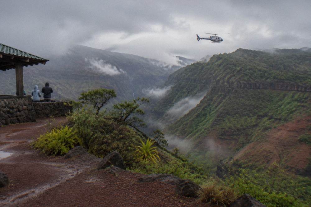 A helicopter hovering above a green canyon rim with mist in the distance