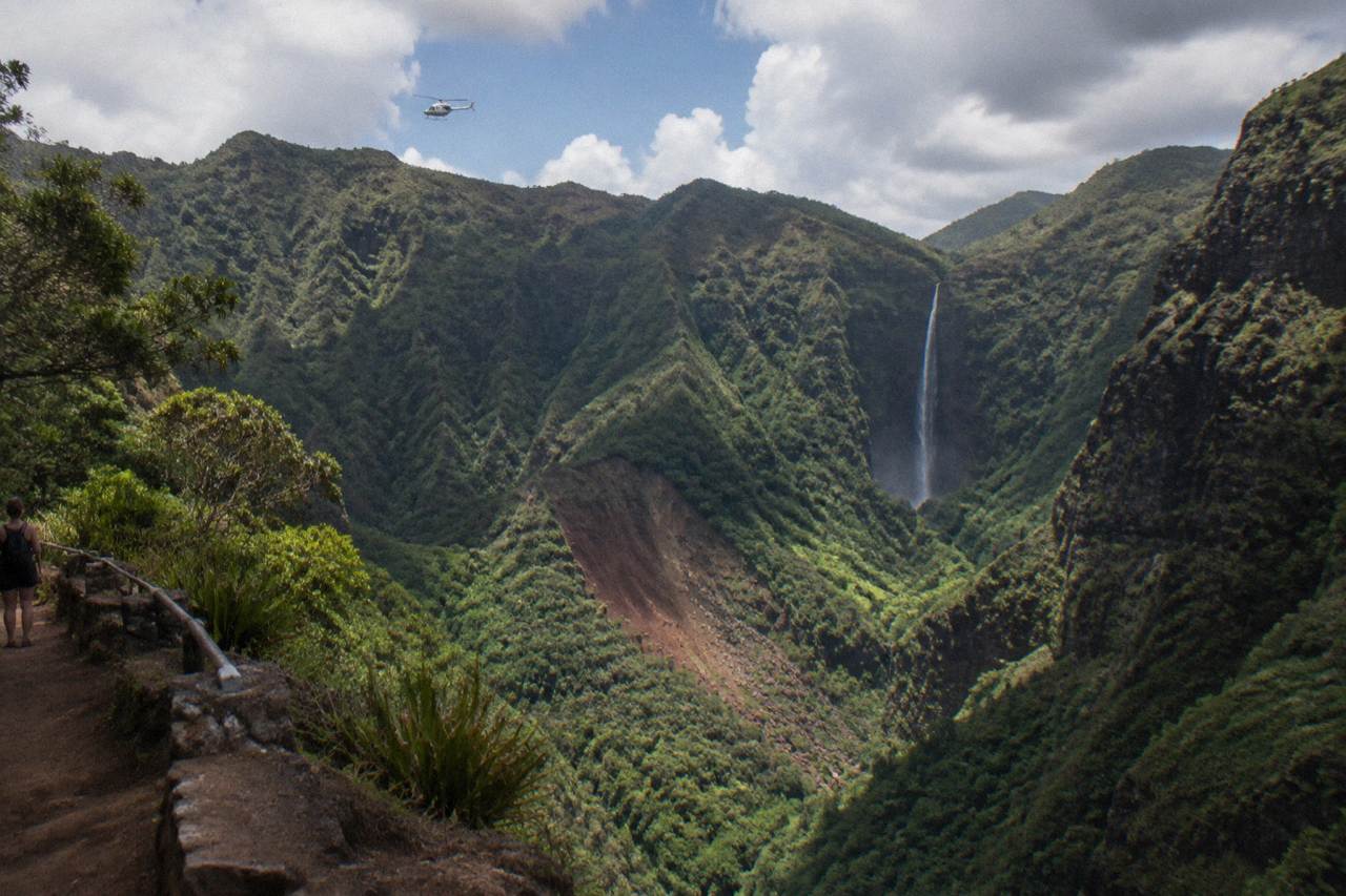 A helicopter flying over deep green Kauai canyons and waterfalls under scattered clouds
