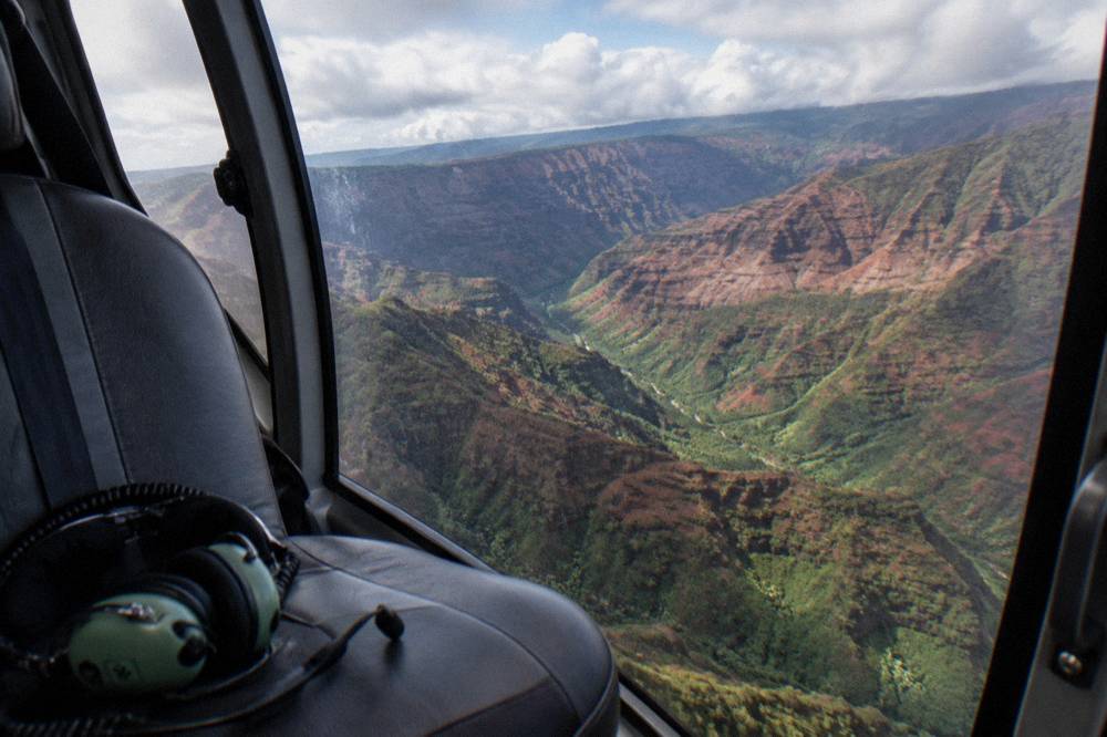 A helicopter cabin view over a deep eroded canyon with red and green ridges under scattered clouds