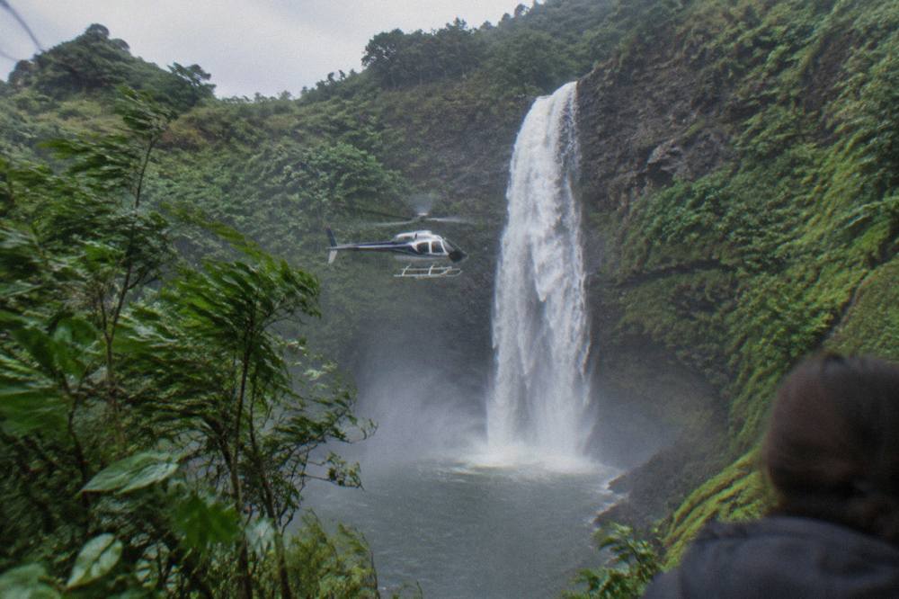 A helicopter hovering near a tall waterfall in a green Kauai valley