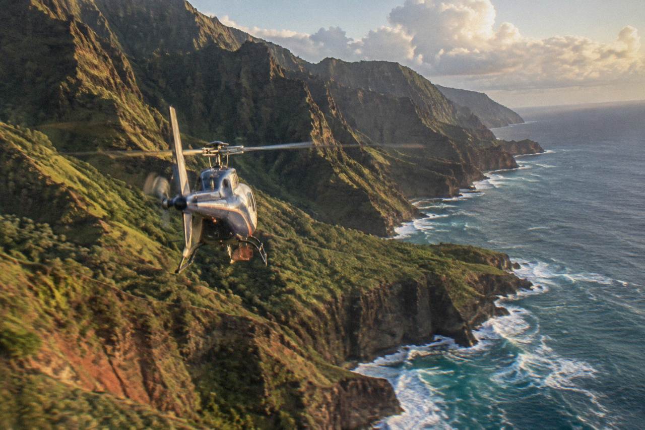 A helicopter flying over green Kauai cliffs and ocean