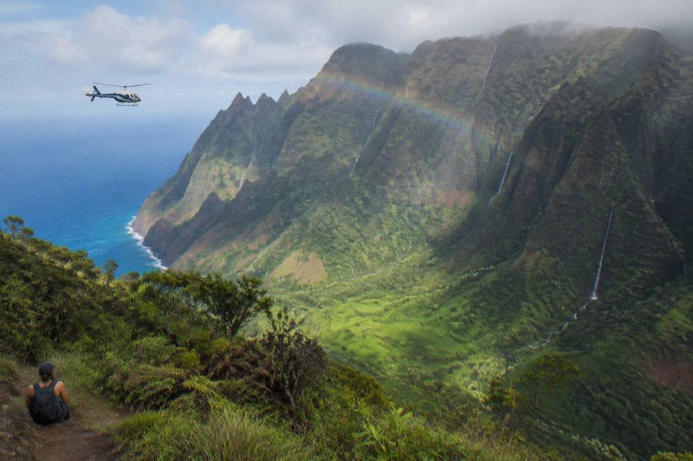 A helicopter flying along towering green sea cliffs with waterfalls streaking the valley walls above the ocean