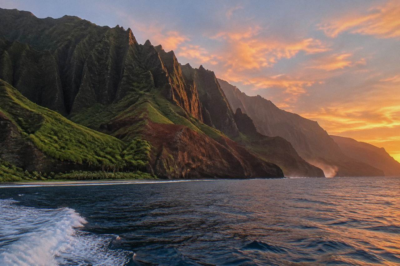 Green sea cliffs of the Na Pali Coast dropping into a blue ocean with wispy clouds over the ridgeline