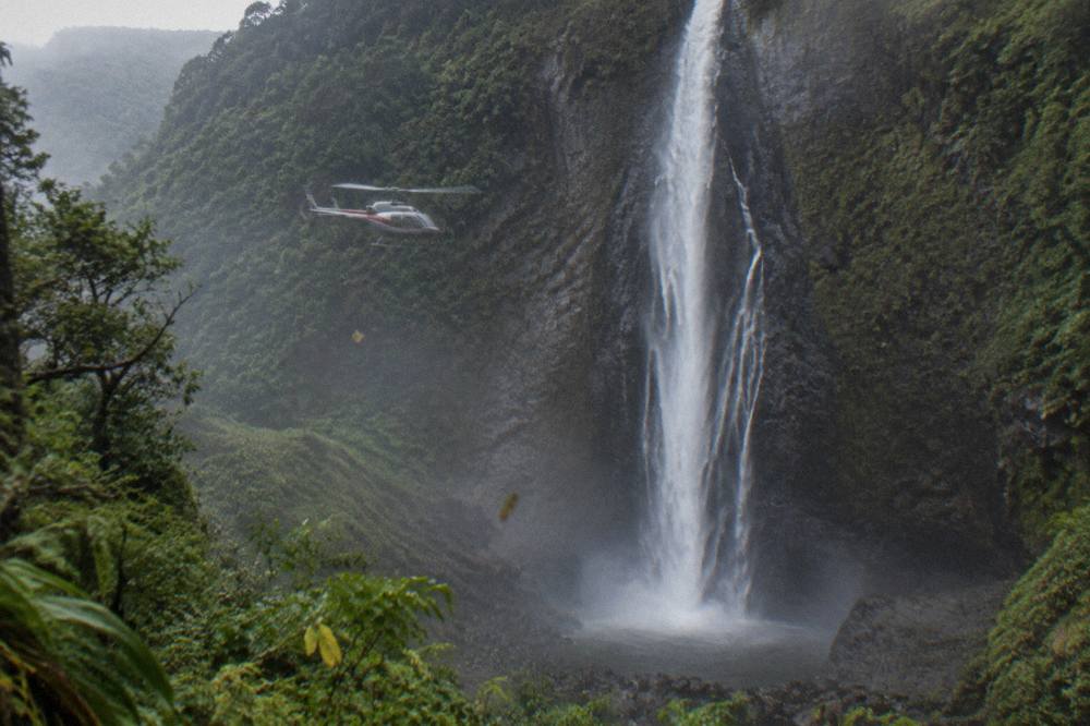 A tour helicopter hovering near a tall ribbon waterfall on a green cliff face
