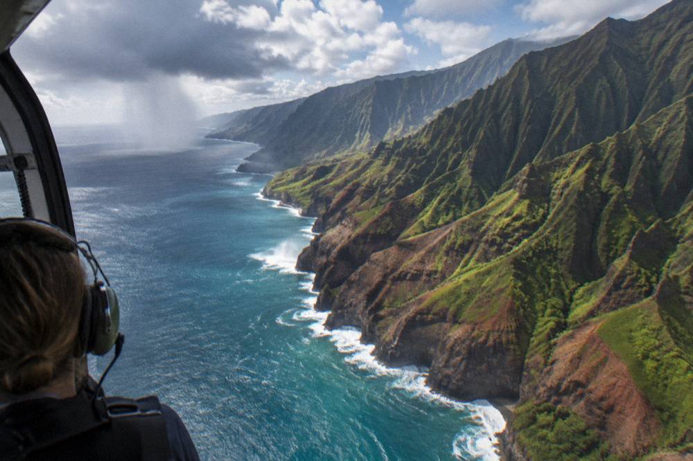 An open-door helicopter in flight above a rugged green coastline