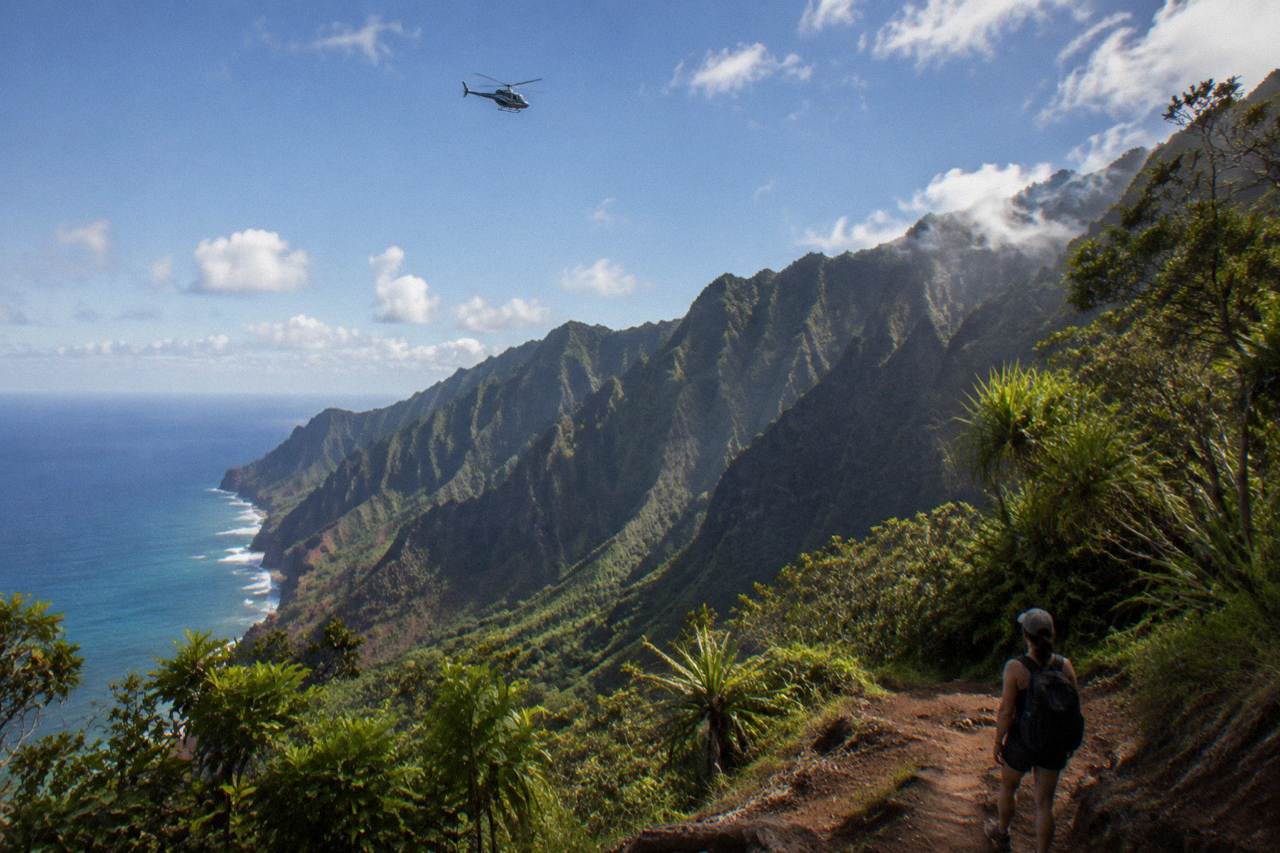 A helicopter flying over green Kauai ridges under scattered clouds