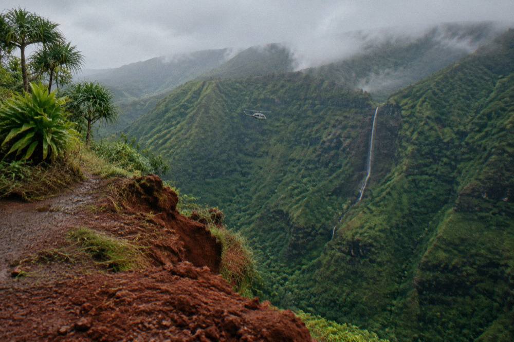 A helicopter flying along a lush green canyon with a thin waterfall visible on the far wall