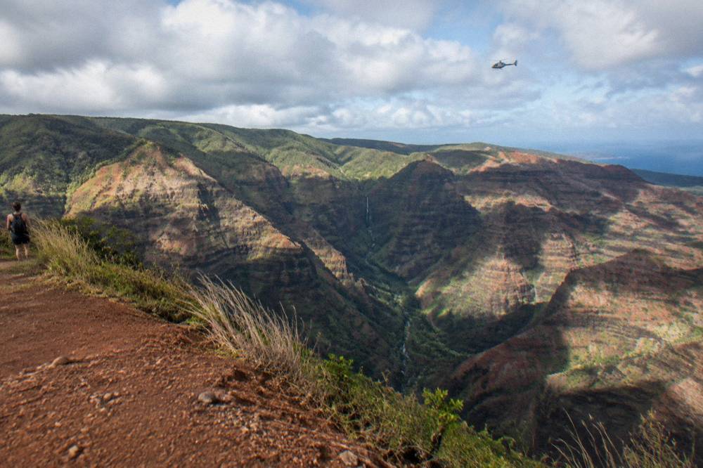 A wide canyon on Kauai with red and green walls and a helicopter flying above the canyon floor