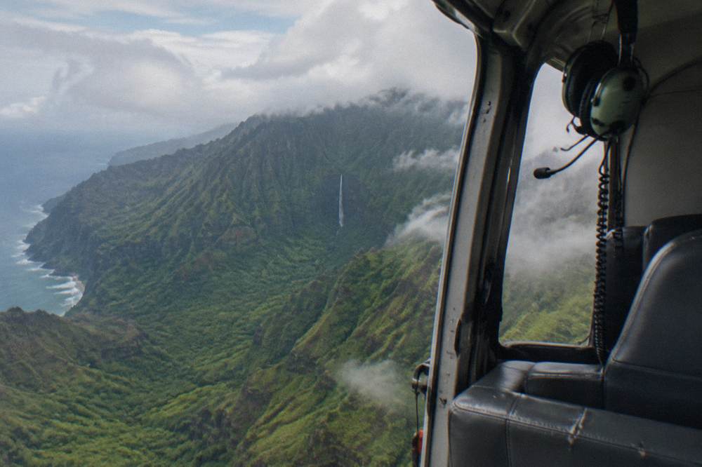 A doors-off helicopter cabin interior above a green valley with a waterfall in the distance