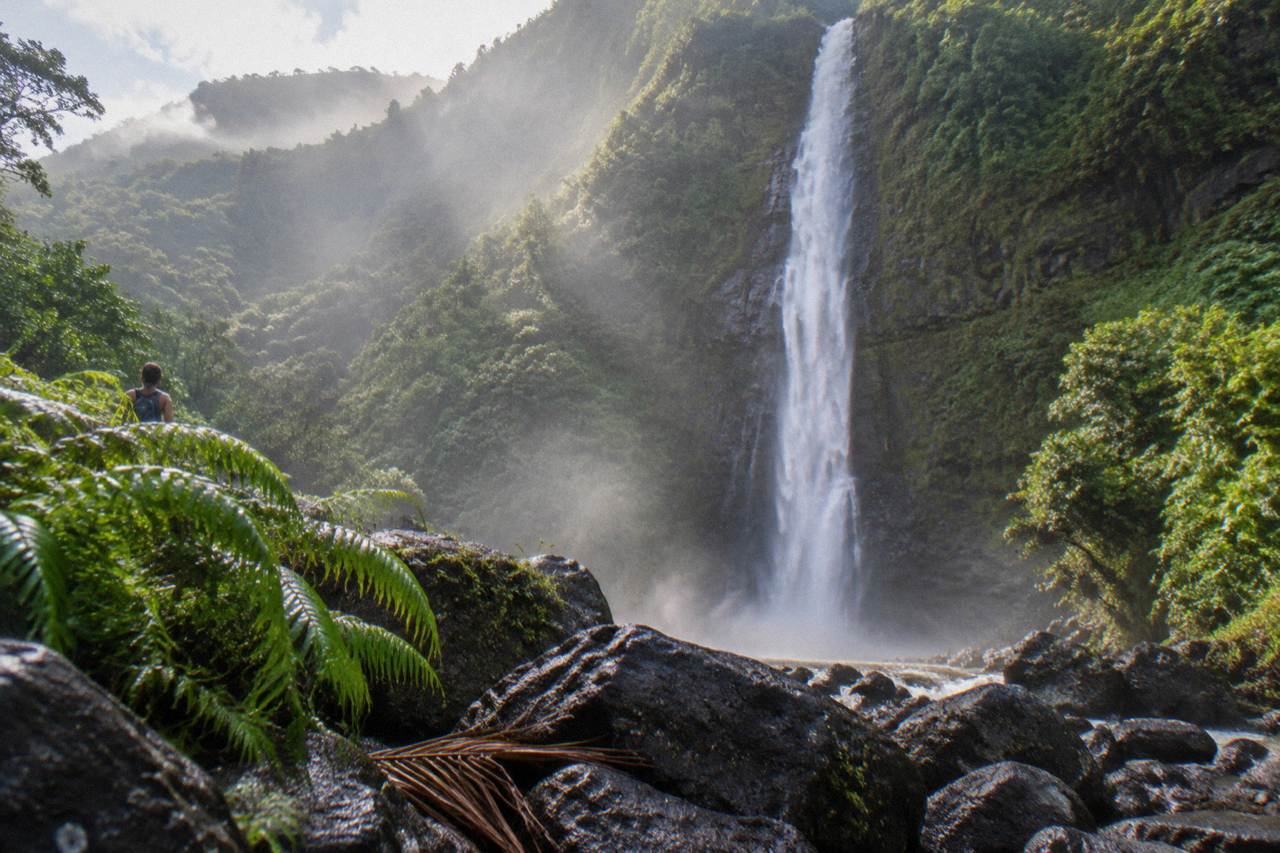 A tall ribbon waterfall dropping down a green cliff face into a jungle valley on Kauai