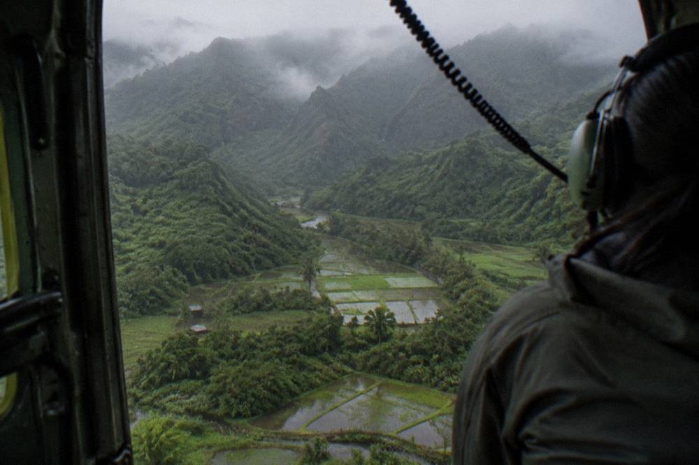 An open-door helicopter in flight above a green valley