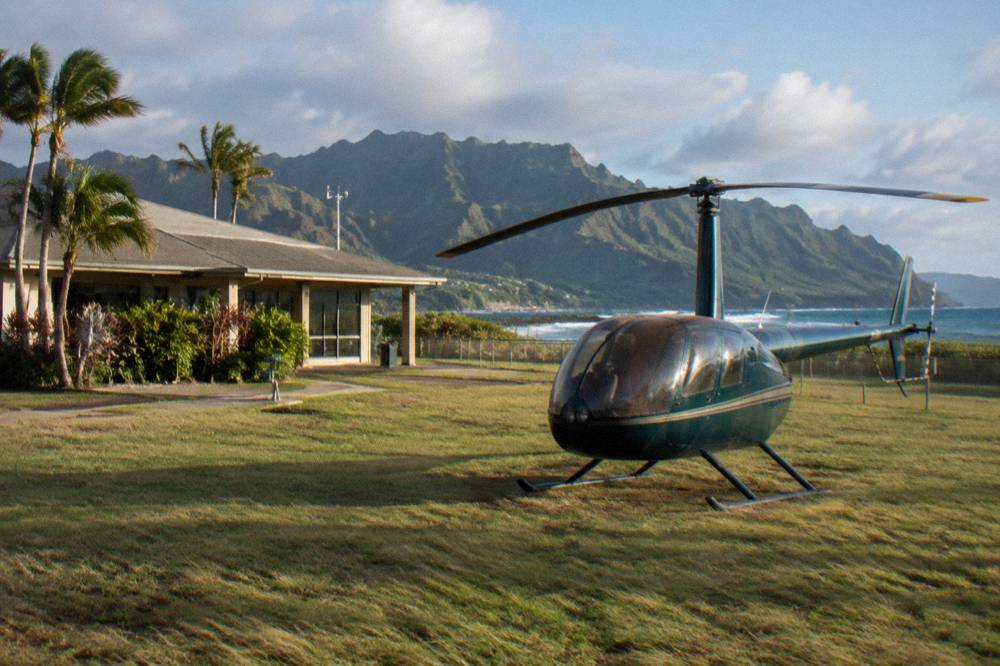 A small helicopter on a grassy apron near the coast with a low-rise terminal building behind it