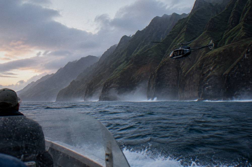 A small helicopter with its doors removed flying along a coastline