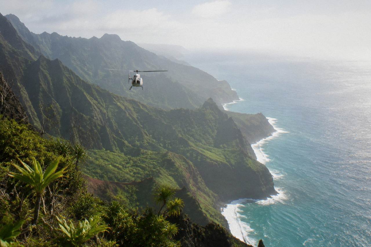 A helicopter flying above green Kauai cliffs with ocean haze on the horizon