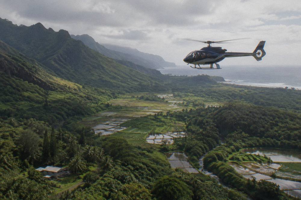 A doors-on helicopter with large windows flying above a green Kauai valley and distant coastline