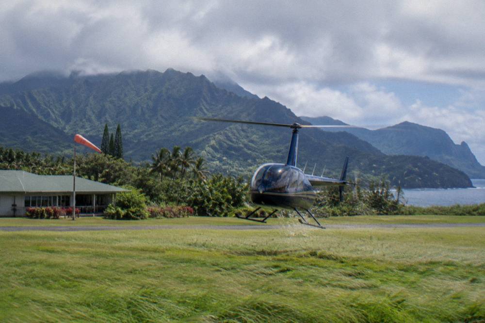 A helicopter lifting off from a grassy heliport with green mountains in the background