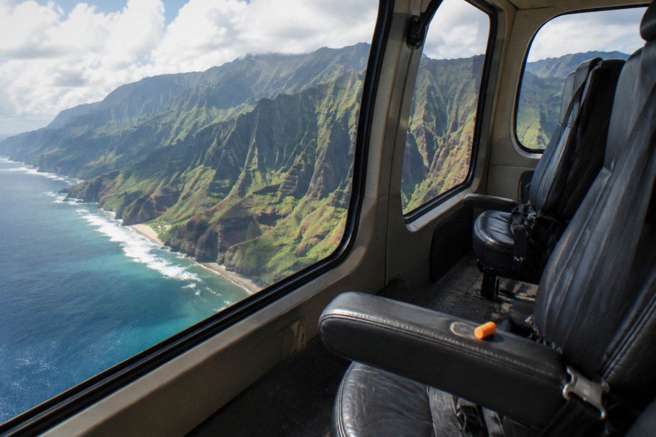 A helicopter cabin interior with empty passenger seats and large window views of green Kauai cliffs