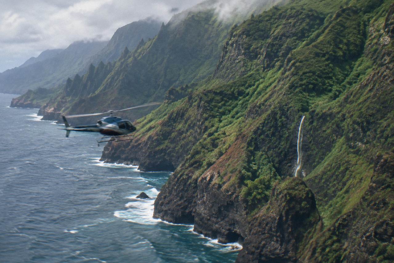 A helicopter flying over green Kauai cliffs above the ocean