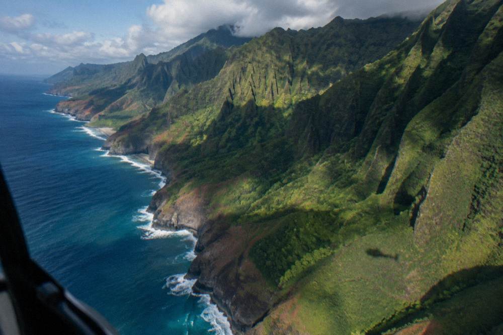 A wide aerial view of a folded coastline with ridges meeting the sea under scattered clouds