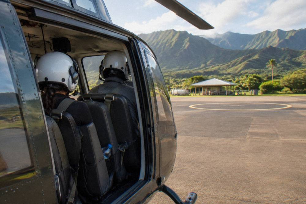 Doors-off private helicopter cabin with two passengers wearing goggles and harnesses, green mountains behind