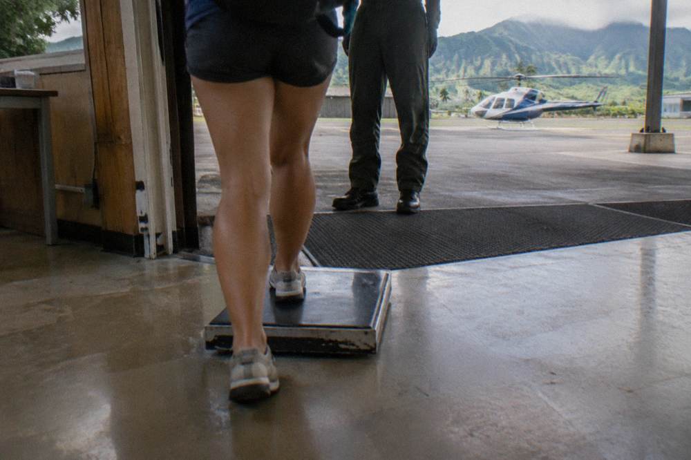 Passenger stepping onto a scale at a helicopter check-in counter with a pilot in the background