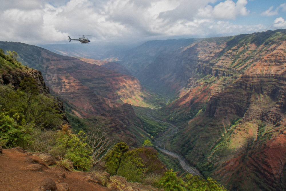 Helicopter flying along a wide red canyon with layered ridge lines under scattered clouds