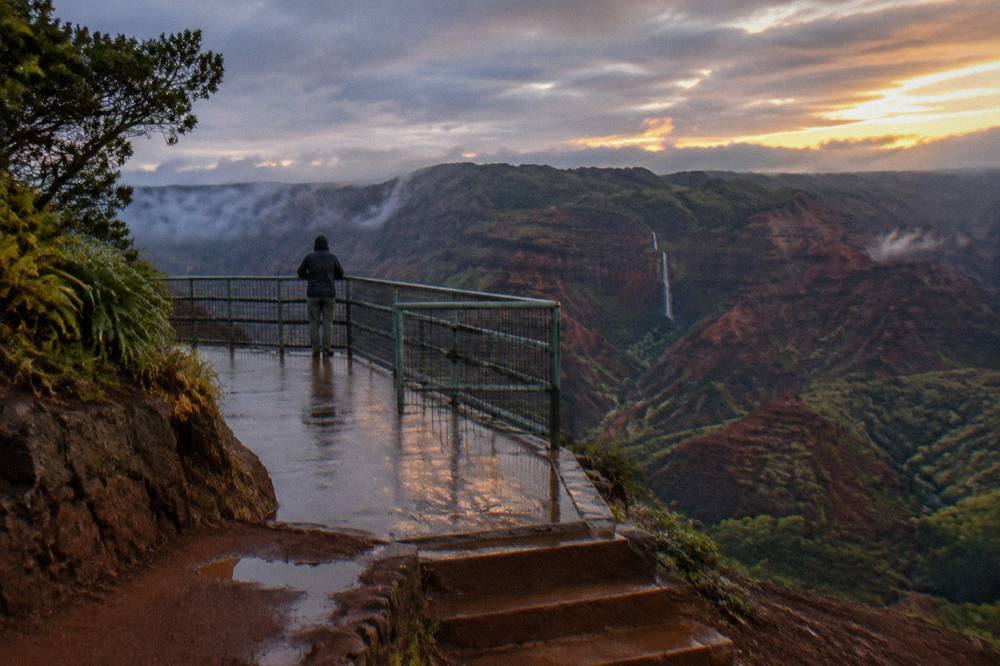 A roadside overlook platform above a wide red canyon with a thin waterfall line on the far wall